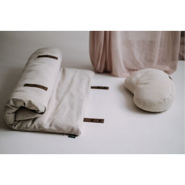 Zestaw do medytacji: zrolowana mata i beżowa poduszka fasolka WellB ustawione na jasnej powierzchni, obok różowej zasłony.